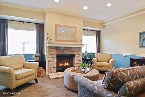 A cozy living room with a stone fireplace in the center, flanked by two windows with dark curtains. The room features three upholstered armchairs and a patterned sofa arranged around a round ottoman with decorative items on top. The walls are painted beige and blue with white trim, and a framed mirror hangs above the fireplace.