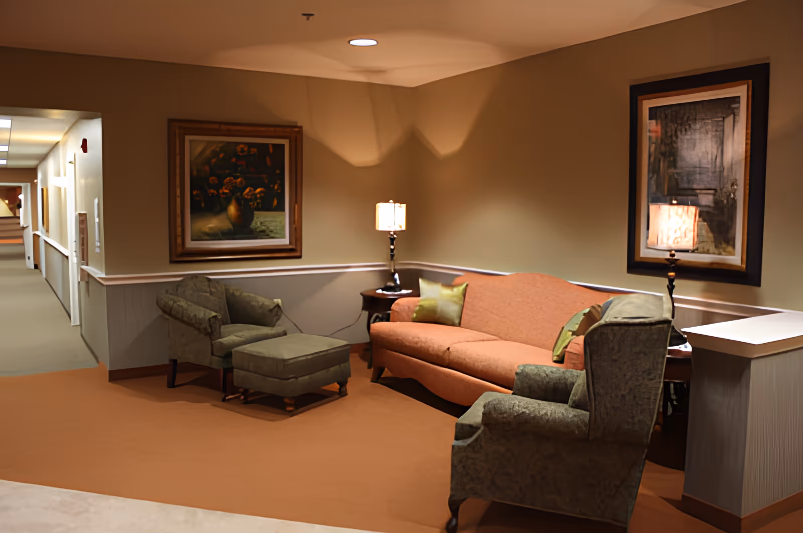 A cozy sitting area in a hallway of an assisted living facility featuring a peach-colored sofa with green pillows, two green upholstered armchairs, and a matching ottoman. The walls are beige with white trim, decorated with framed paintings. Two table lamps provide warm lighting on side tables beside the sofa.