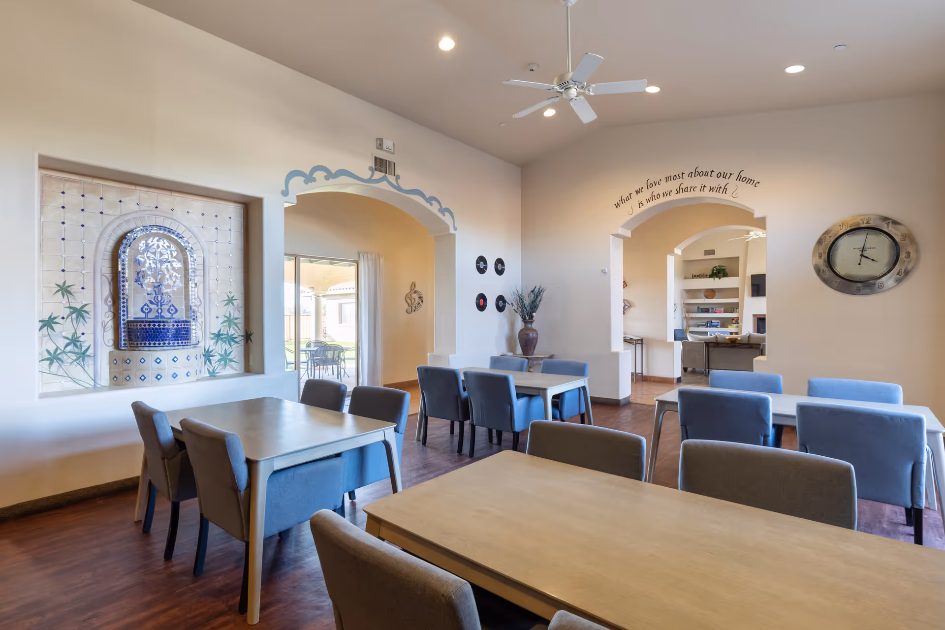 A bright dining room in BridgeWater Assisted Living - Tucson featuring several tables with blue upholstered chairs. The room has wooden flooring, a decorative wall fountain, and arched doorways leading to other areas. A wall clock and a large vase with plants are visible, along with a ceiling fan and recessed lighting. A quote on the wall reads, 'What we love most about our home is who we share it with.'