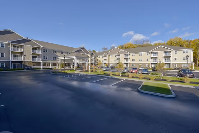 Exterior view of a large, multi-story senior living facility with beige siding and stone accents, surrounded by a parking lot with several cars and landscaped greenery under a clear blue sky.