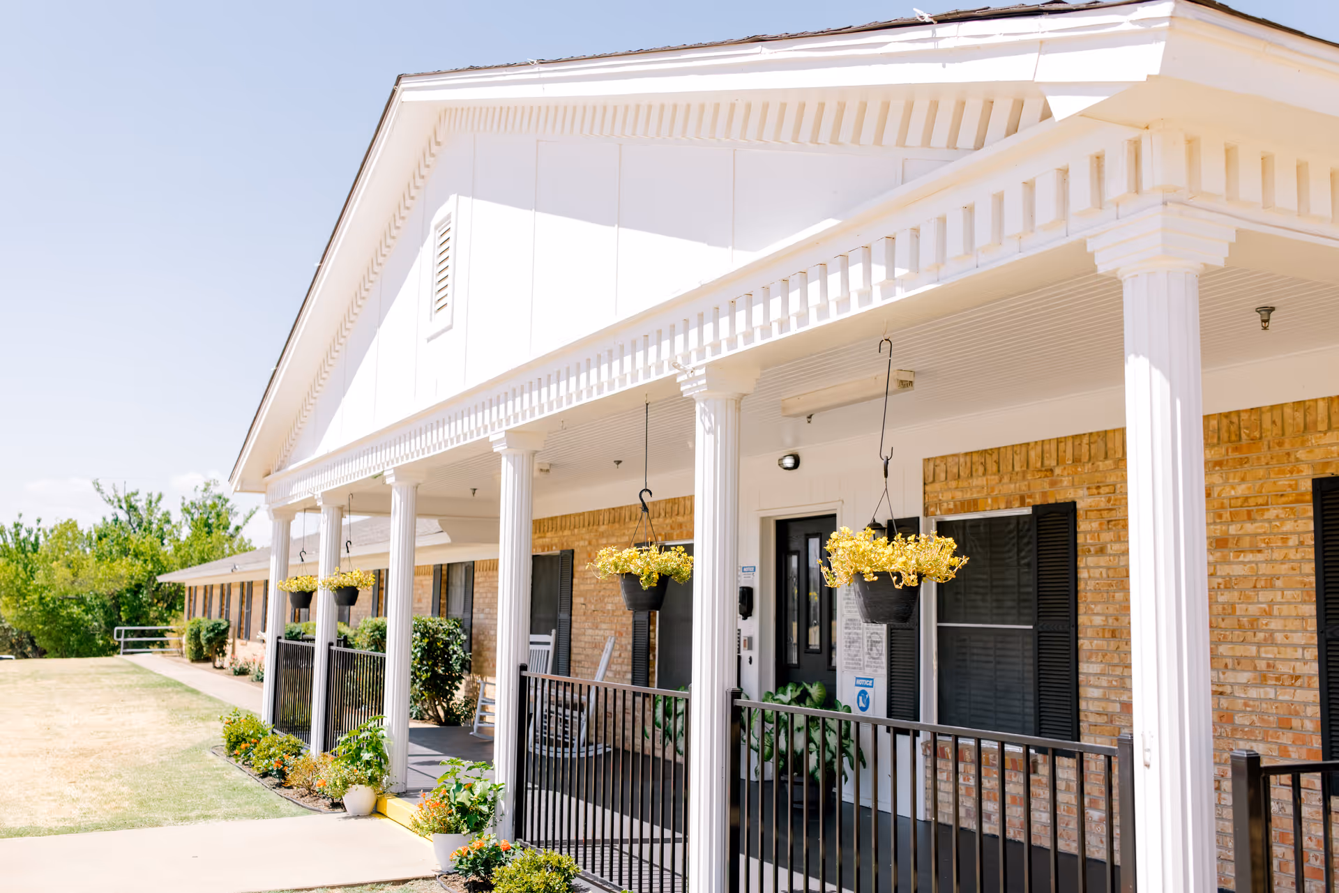 Exterior view of Keeneland Nursing & Rehabilitation showing a long porch with white columns, hanging flower pots with yellow flowers, black railings, and brick walls with black window shutters under a clear blue sky.
