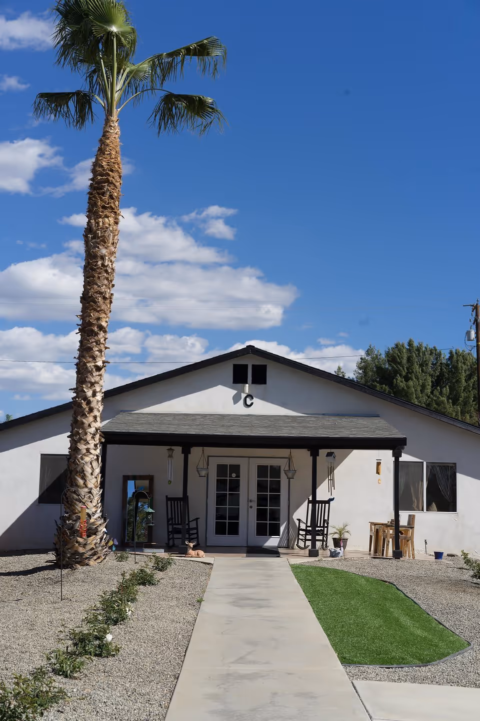 Exterior view of a single-story building with a covered porch featuring two rocking chairs and hanging lanterns. A concrete pathway leads to the entrance with double glass doors. There is a tall palm tree on the left side and a small patch of green grass on the right side of the pathway. The sky is clear with some clouds.