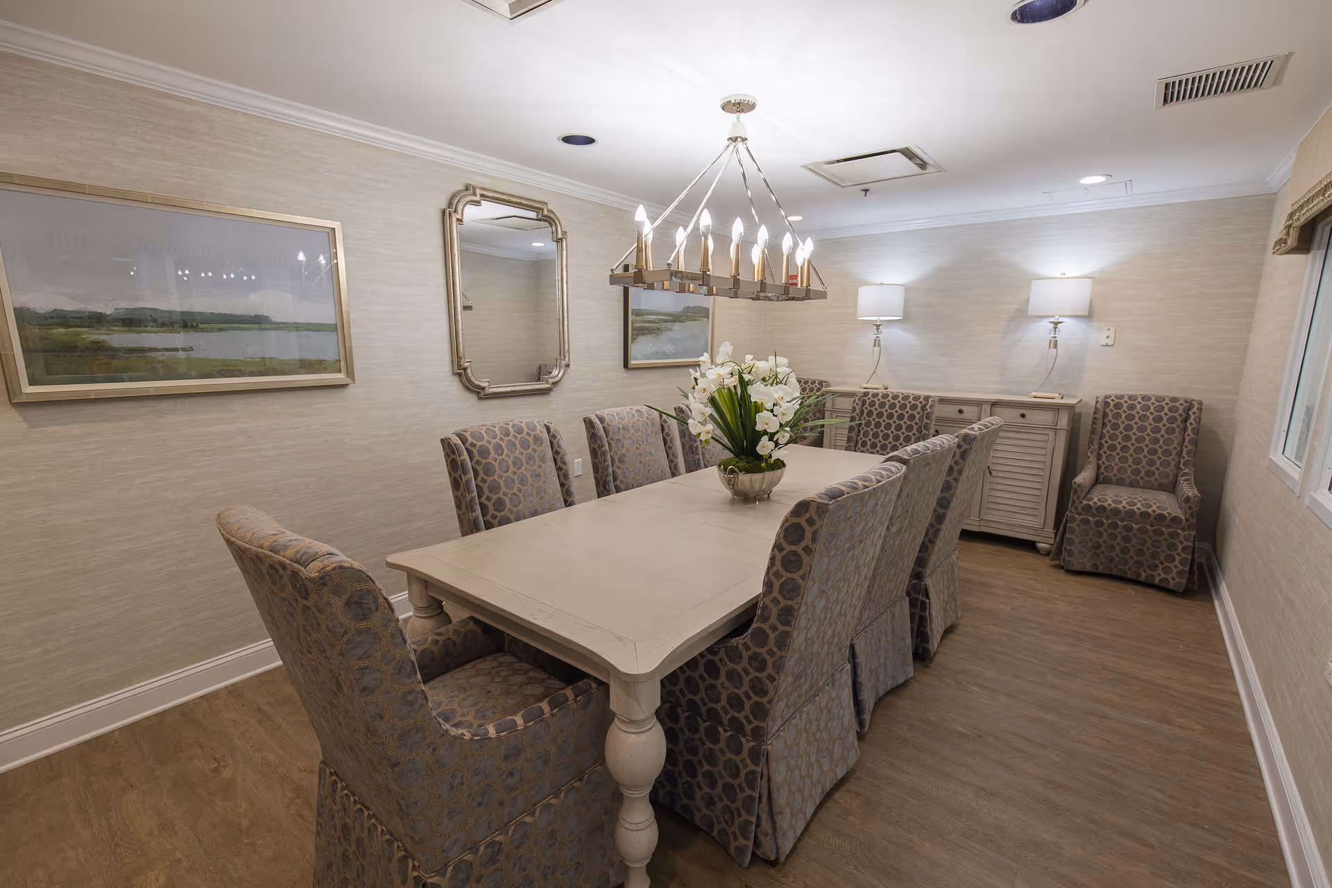 A well-lit dining room with a long rectangular wooden table surrounded by eight upholstered chairs with a patterned fabric. A modern chandelier with multiple bulbs hangs above the table. The room has light-colored walls with two framed landscape paintings and a decorative mirror. There is a sideboard with two table lamps and two additional chairs in the corner. The floor is wood, and there is a window on the right side.