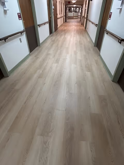 A long, empty hallway in a senior living facility with light wood flooring, handrails on both sides, and several closed doors along the walls. The hallway is well-lit with ceiling lights and has a clean, orderly appearance.