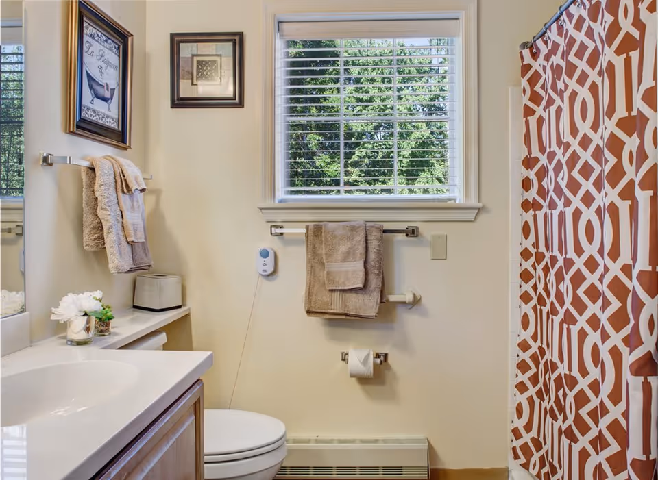 A clean bathroom with a white sink and wooden cabinet, a toilet, beige towels hanging on towel racks, a window with white blinds showing green trees outside, framed artwork on the wall, and a shower with a red and white patterned curtain.
