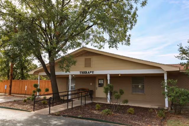 Exterior view of a single-story brick building with a beige roof and a sign above the entrance that reads 'THERAPY'. There is a wheelchair ramp leading to the door, surrounded by small shrubs and trees, under a partly cloudy sky.