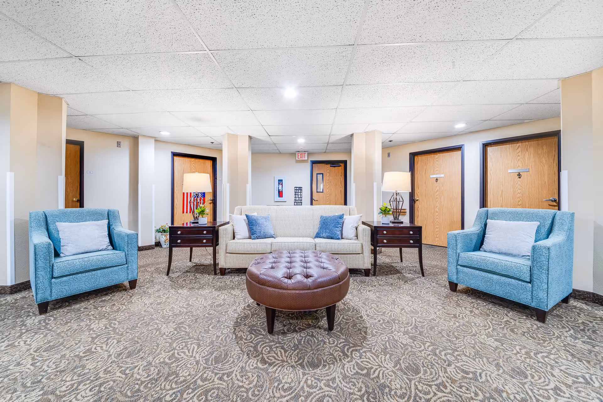 A seating area in a senior living facility with a beige sofa flanked by two dark wooden side tables with lamps and plants, two blue armchairs with light-colored pillows, and a round brown tufted ottoman in the center. The background shows beige walls, wooden doors, and a patterned carpet.