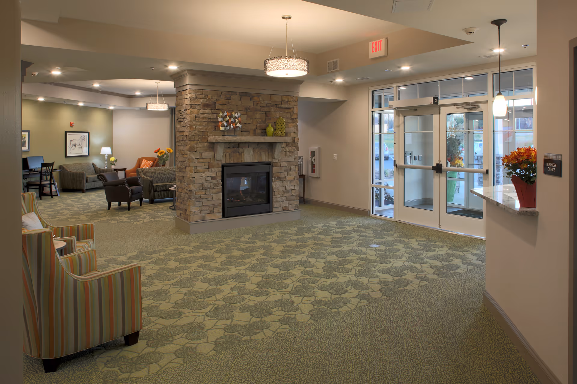 Lobby of a senior living community with seating, a stone fireplace, patterned carpet, and glass entrance doors.