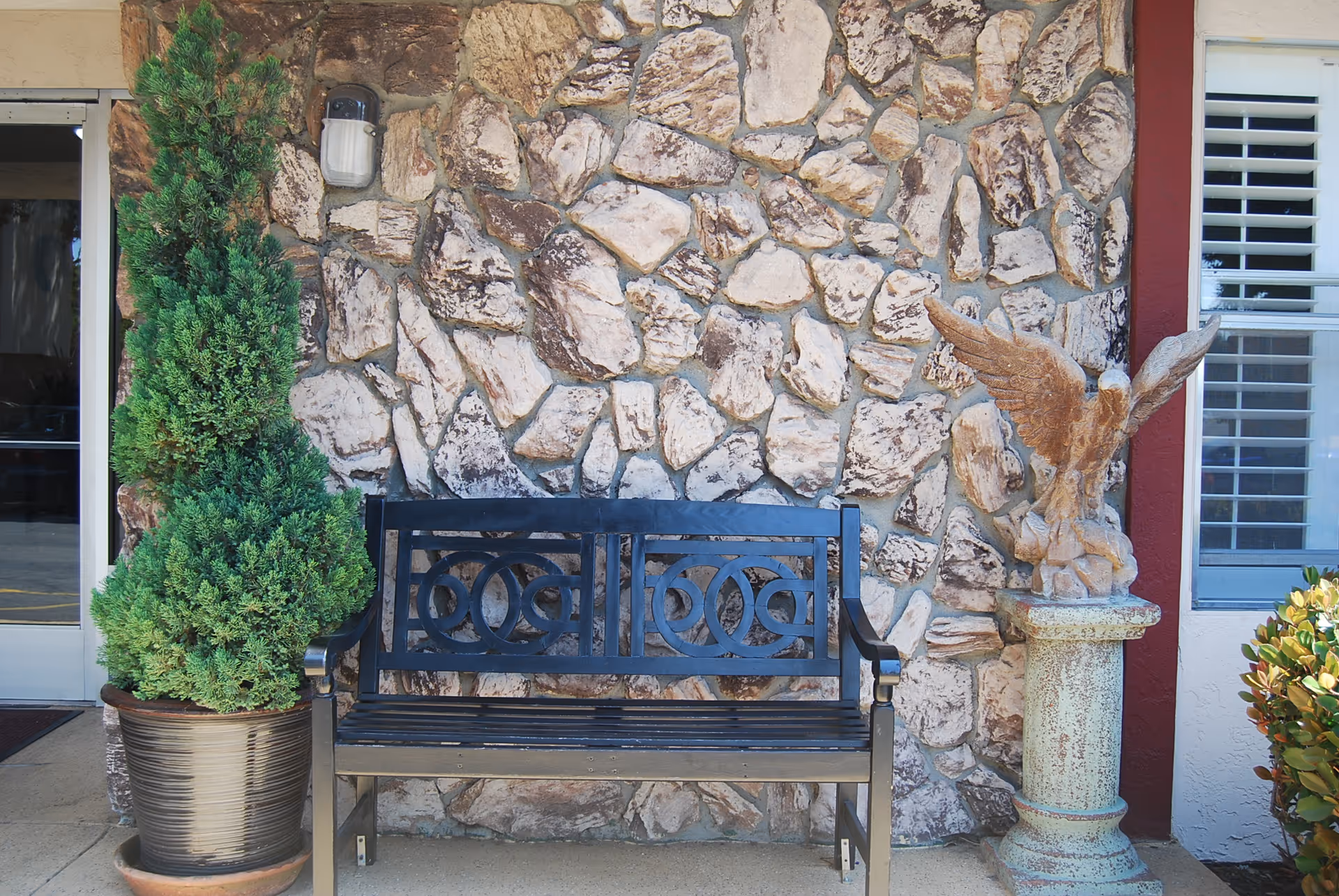 Black metal bench placed against a stone wall with a potted green shrub on the left and a stone eagle statue on a pedestal on the right, outside a building entrance with a glass door and window with white shutters.