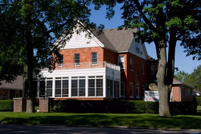 Brick two-story building with a large glassed-in sunroom, set behind trees and a manicured lawn.
