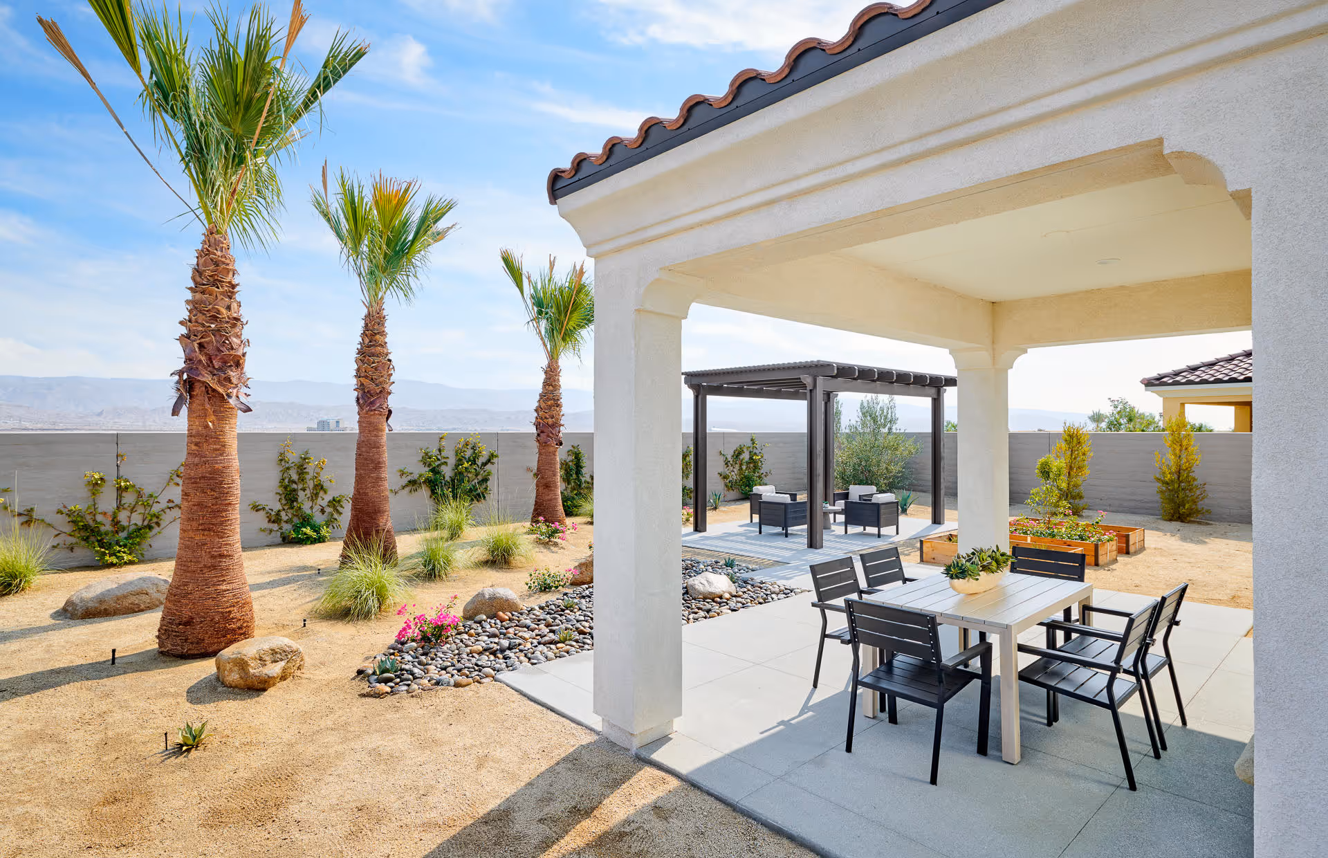 Outdoor patio area with a covered seating space featuring a table and four chairs. In the background, there is a pergola with additional seating. The area is landscaped with palm trees, rocks, and small plants, enclosed by a gray wall with mountains visible in the distance under a blue sky.