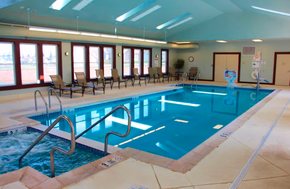 Indoor swimming pool area with clear blue water, surrounded by beige tiled flooring. There are several lounge chairs lined up along the windows on one side. The room has large windows letting in natural light and overhead fluorescent lighting. A small hot tub is visible in the foreground with metal handrails.