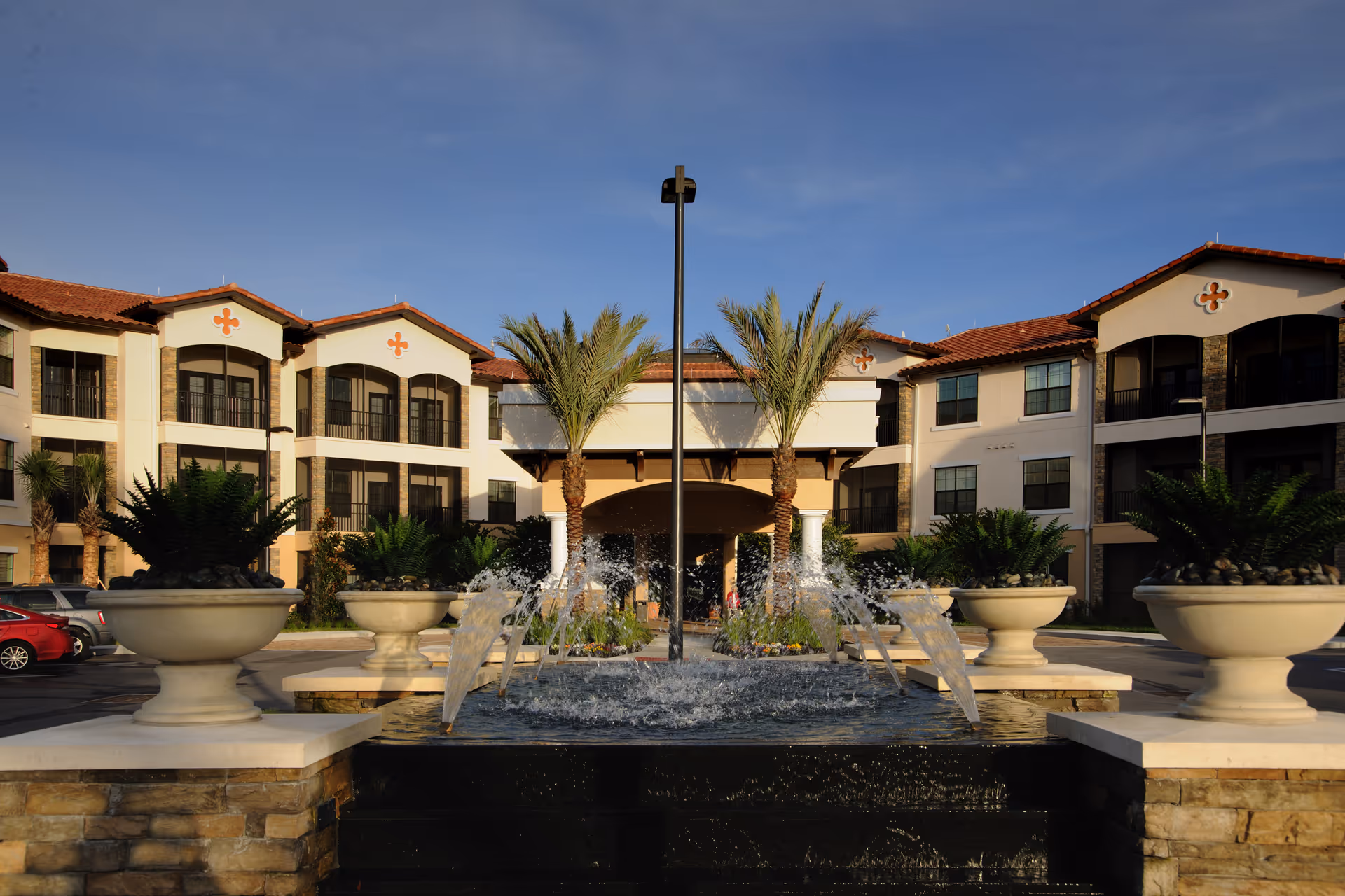 Exterior view of Allegro Senior Living facility in Winter Park, FL, featuring a central water fountain with multiple streams of water, large potted plants, palm trees, and a multi-story building with balconies and red-tiled roofs under a clear blue sky.