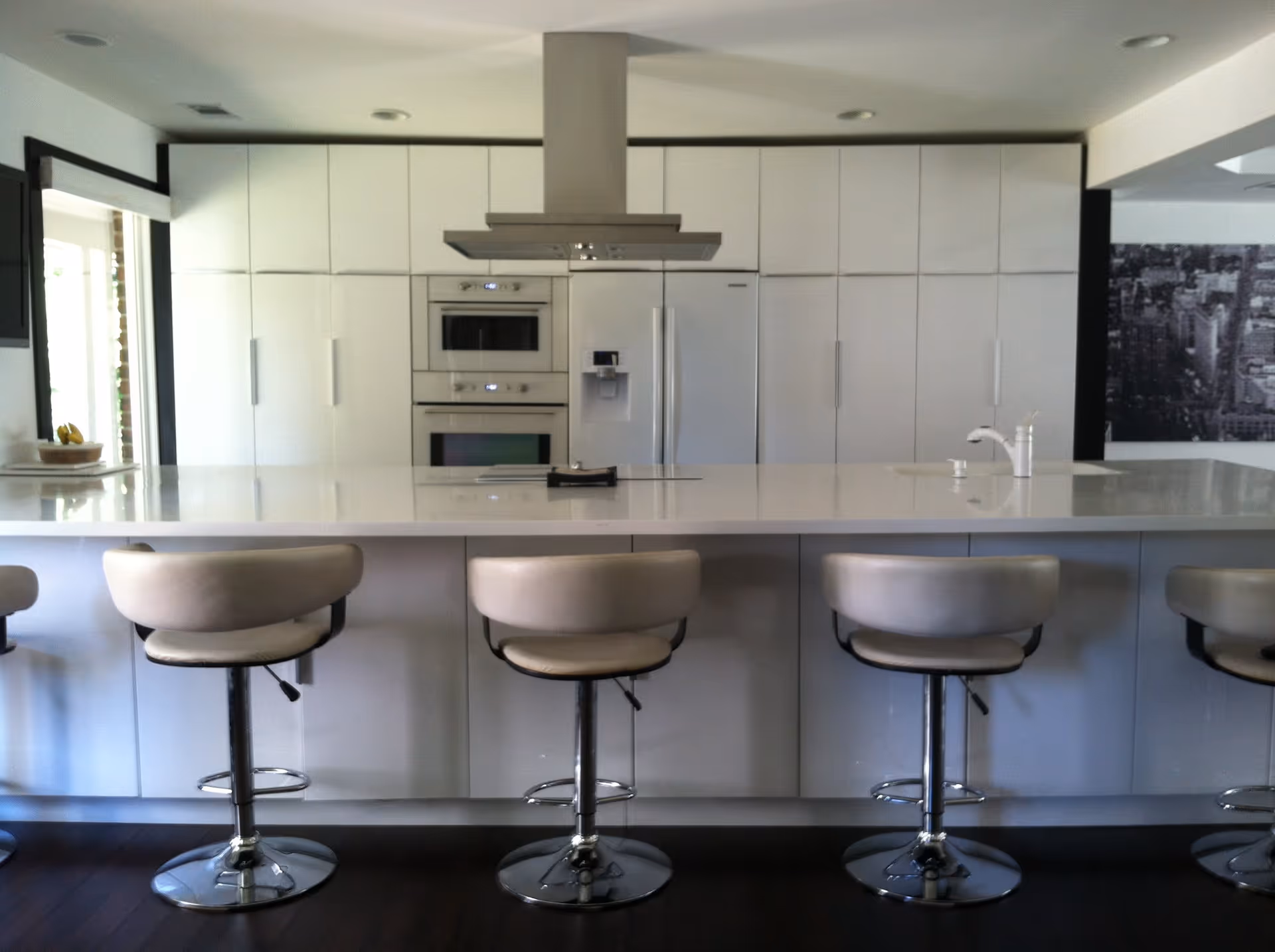 Modern kitchen with a large white island countertop and four beige bar stools with chrome bases. The back wall features white cabinetry, a built-in oven and microwave, a refrigerator with a water dispenser, and a stainless steel range hood. The floor is dark wood, and there is a black and white cityscape picture on the right wall.