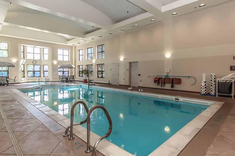 Bright indoor swimming pool with handrails, lounge chairs, tables, and large windows in a senior living facility.