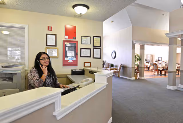 A woman with long dark hair and glasses is sitting behind a reception desk, talking on the phone. The reception area has beige walls with framed certificates and a red fire alarm box on the wall. In the background, there is a spacious common area with tables and chairs, and large windows letting in natural light.