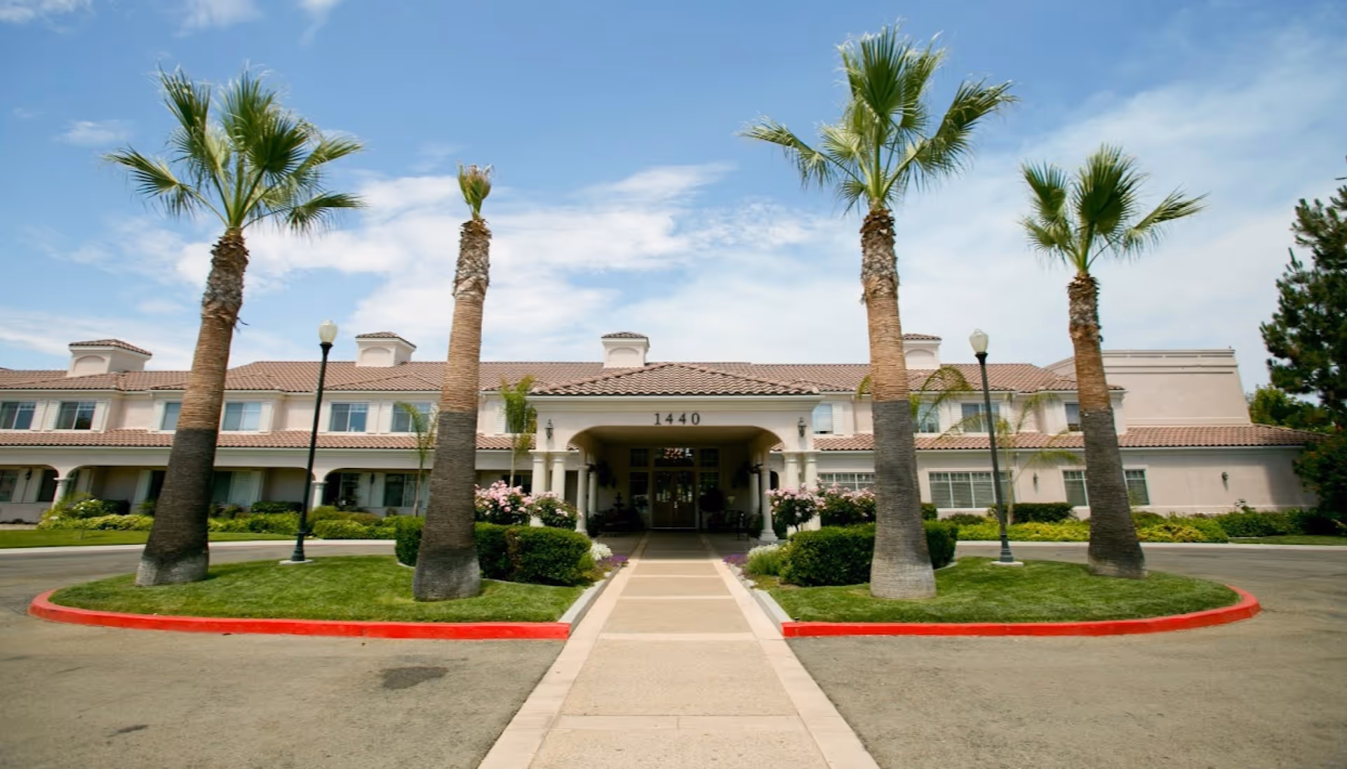 Front exterior view of a senior living facility named Aegis Living Shadowridge with a driveway leading to the entrance, lined with palm trees and lamp posts under a partly cloudy sky.