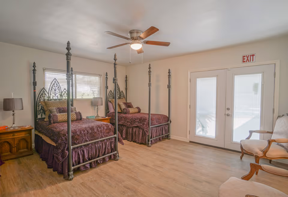 A bedroom with two ornate four-poster twin beds dressed in purple bedding. Each bed has decorative pillows and is flanked by wooden nightstands with lamps. There is a ceiling fan above, a window with blinds behind the beds, and double glass doors with an exit sign above them. Two upholstered chairs are positioned near the doors on a wooden floor.