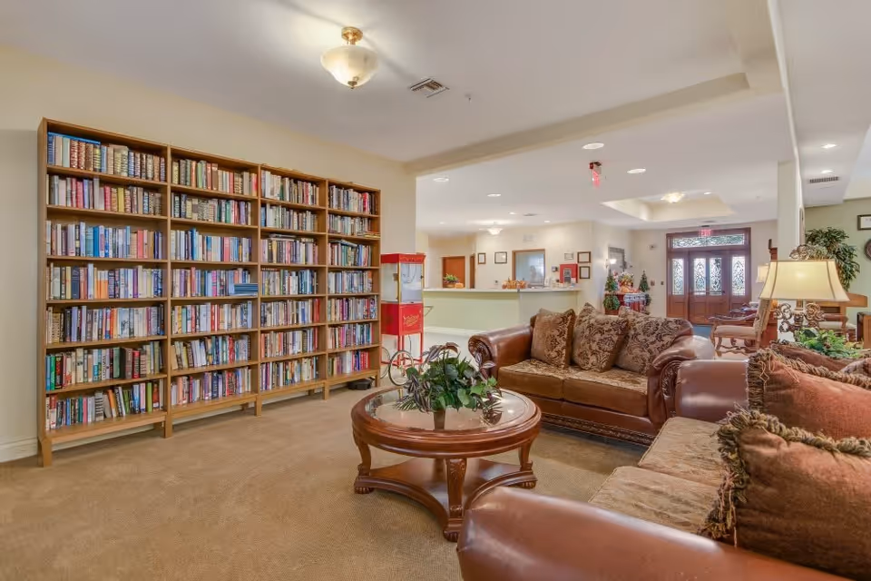 A cozy living room area with brown leather sofas adorned with patterned cushions, a round wooden coffee table with a plant centerpiece, and a large wooden bookshelf filled with books. In the background, there is a reception desk and a wooden door with glass panels, along with warm lighting fixtures on the ceiling.