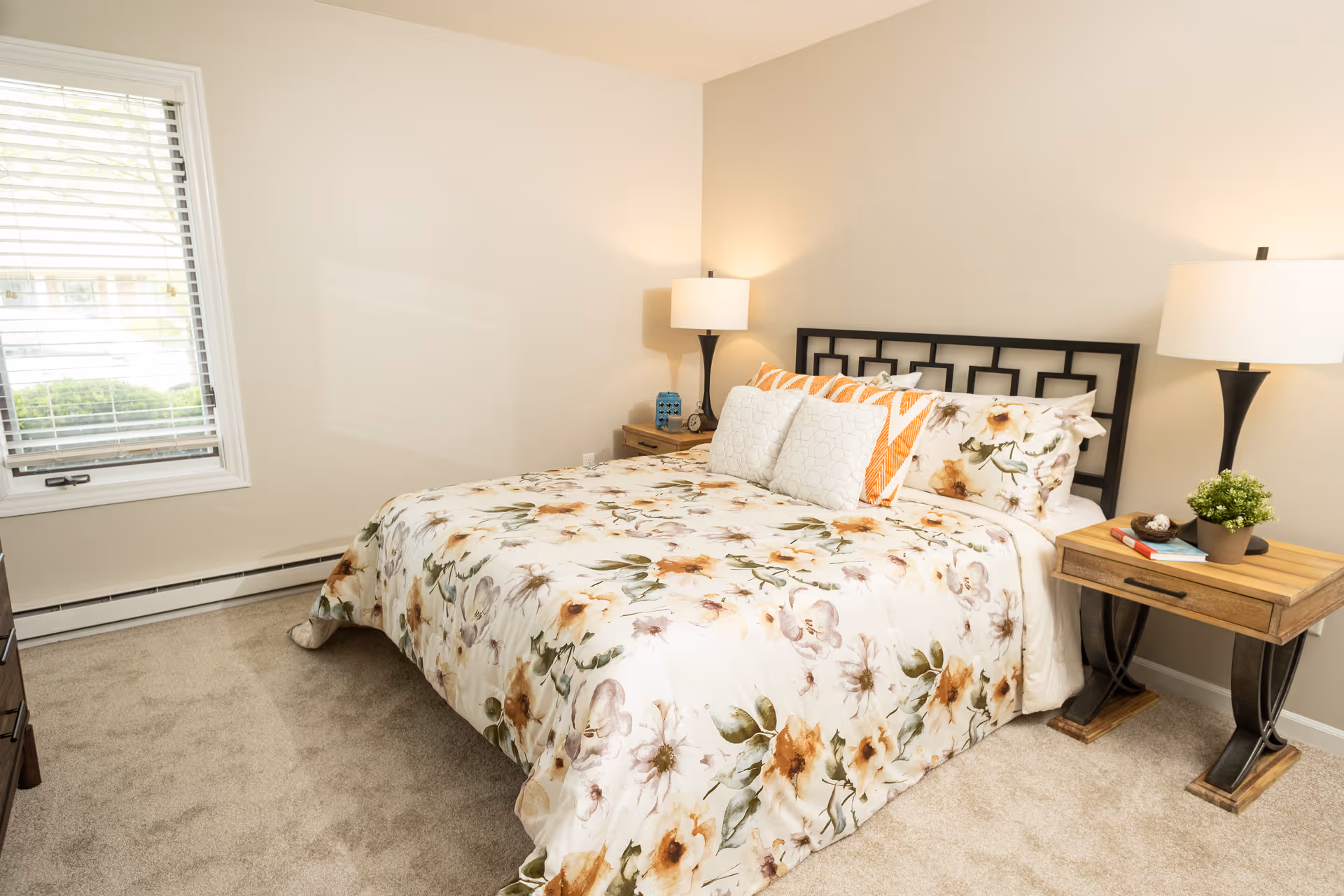 Well-lit bedroom with a floral bedspread, two nightstands with lamps, and a window with blinds.