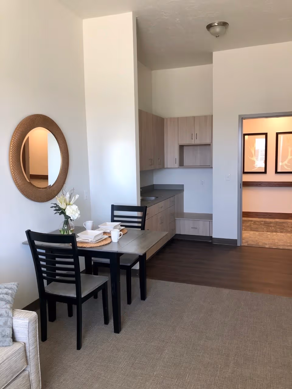 A small dining area with a table set for two, featuring black chairs and a vase with white flowers. The area is adjacent to a kitchenette with light wood cabinets and a gray countertop. A round decorative mirror hangs on the wall above the dining table. The floor is carpeted near the dining area and transitions to wood flooring near the kitchenette. An open doorway leads to another room with framed artwork on the walls.