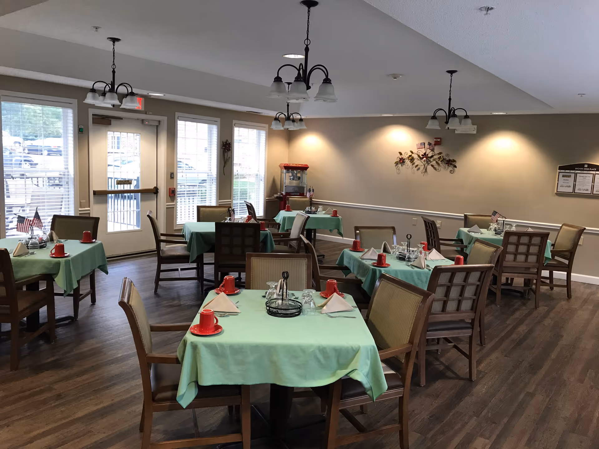 A dining room with several tables covered in green tablecloths, each set with red cups, napkins, and glassware. Wooden chairs surround the tables. The room has large windows and a door letting in natural light. There are ceiling lights and wall decorations, including a popcorn machine in the corner.