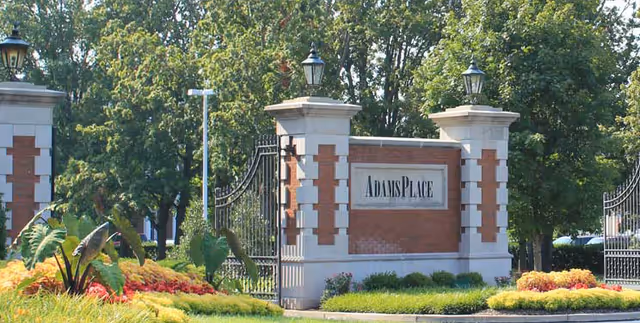 Entrance sign for AdamsPlace facility with brick pillars, black wrought iron gates, and surrounding landscaped greenery including bushes and trees.