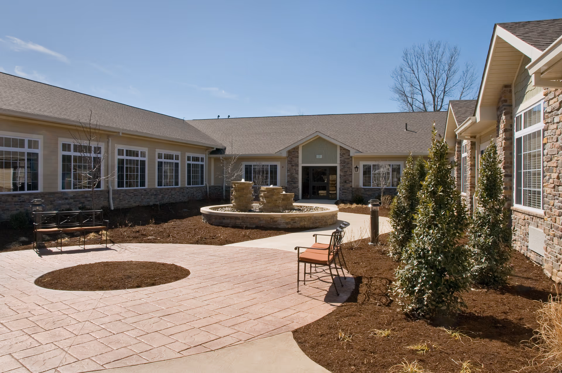 Outdoor courtyard area of a senior living facility with a circular stone fountain in the center, surrounded by a paved walkway. There are benches with orange cushions and some small trees and shrubs planted in mulched beds. The building has stone and siding exterior with multiple windows and a clear blue sky above.