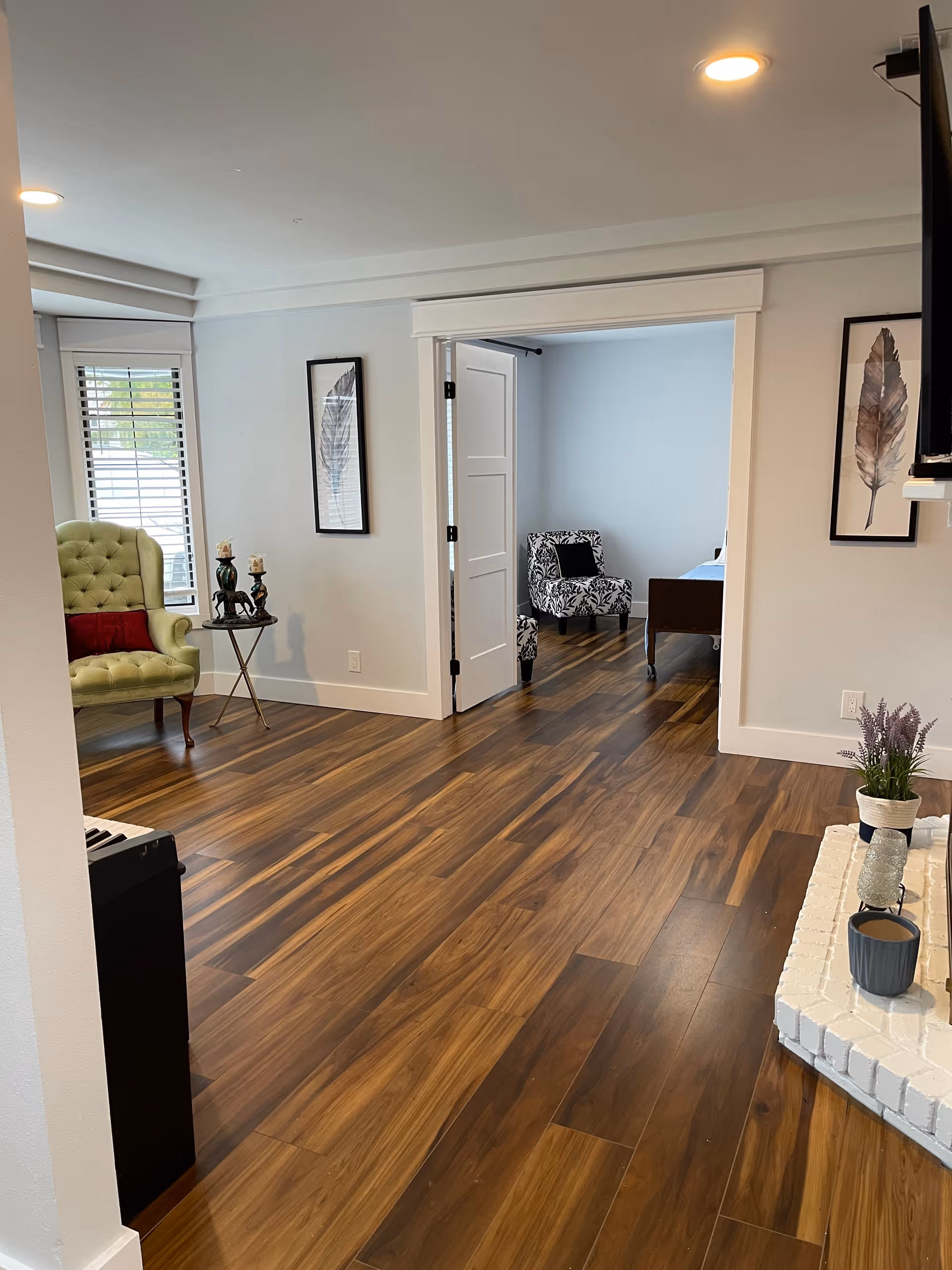 A bright interior room with wooden flooring featuring a green tufted armchair with a red cushion near a window with white blinds. There is a small round side table with decorative items next to the armchair. The room opens into another room with a patterned armchair and a bed. The walls are light-colored with framed feather artwork. A white brick hearth with a potted plant and a cup is visible in the foreground.