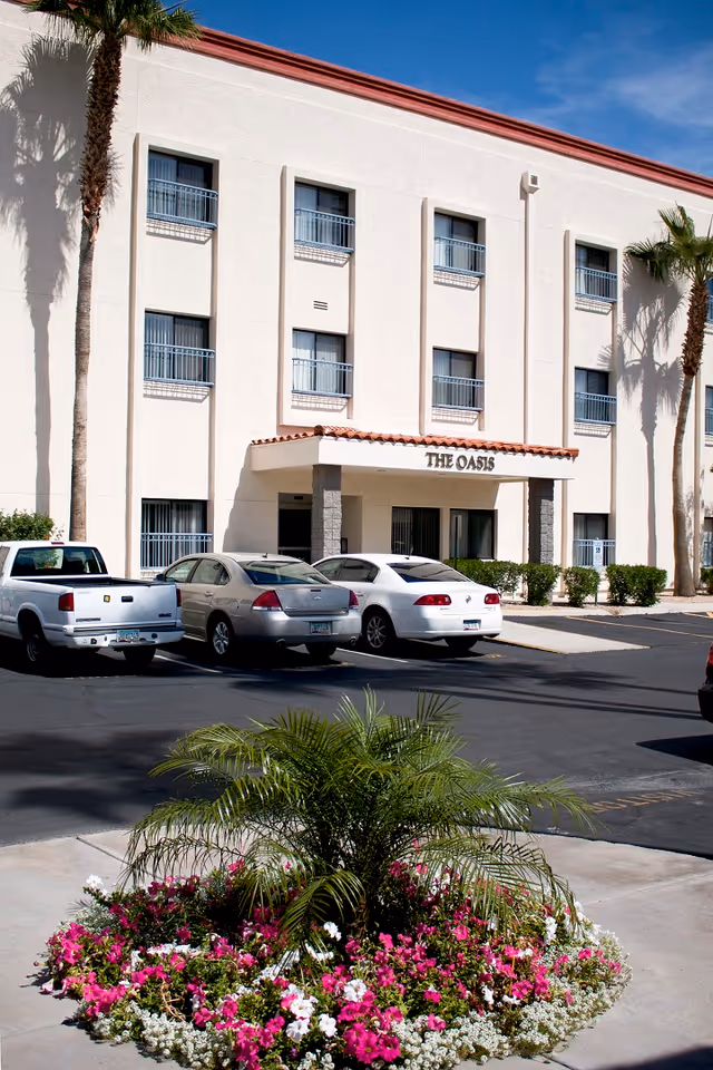 Front entrance of a beige three-story building labeled 'The Oasis' with parked cars and a flowerbed in the foreground.
