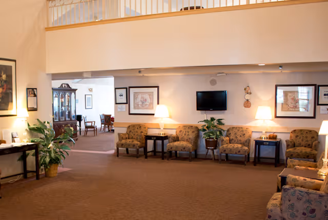 A spacious living room area in a senior living facility with several upholstered chairs arranged along the walls, two side tables with lamps, framed artwork on the walls, a flat-screen TV mounted on the wall, and potted plants. The room has a carpeted floor and an open view into an adjacent dining area with wooden chairs and tables.