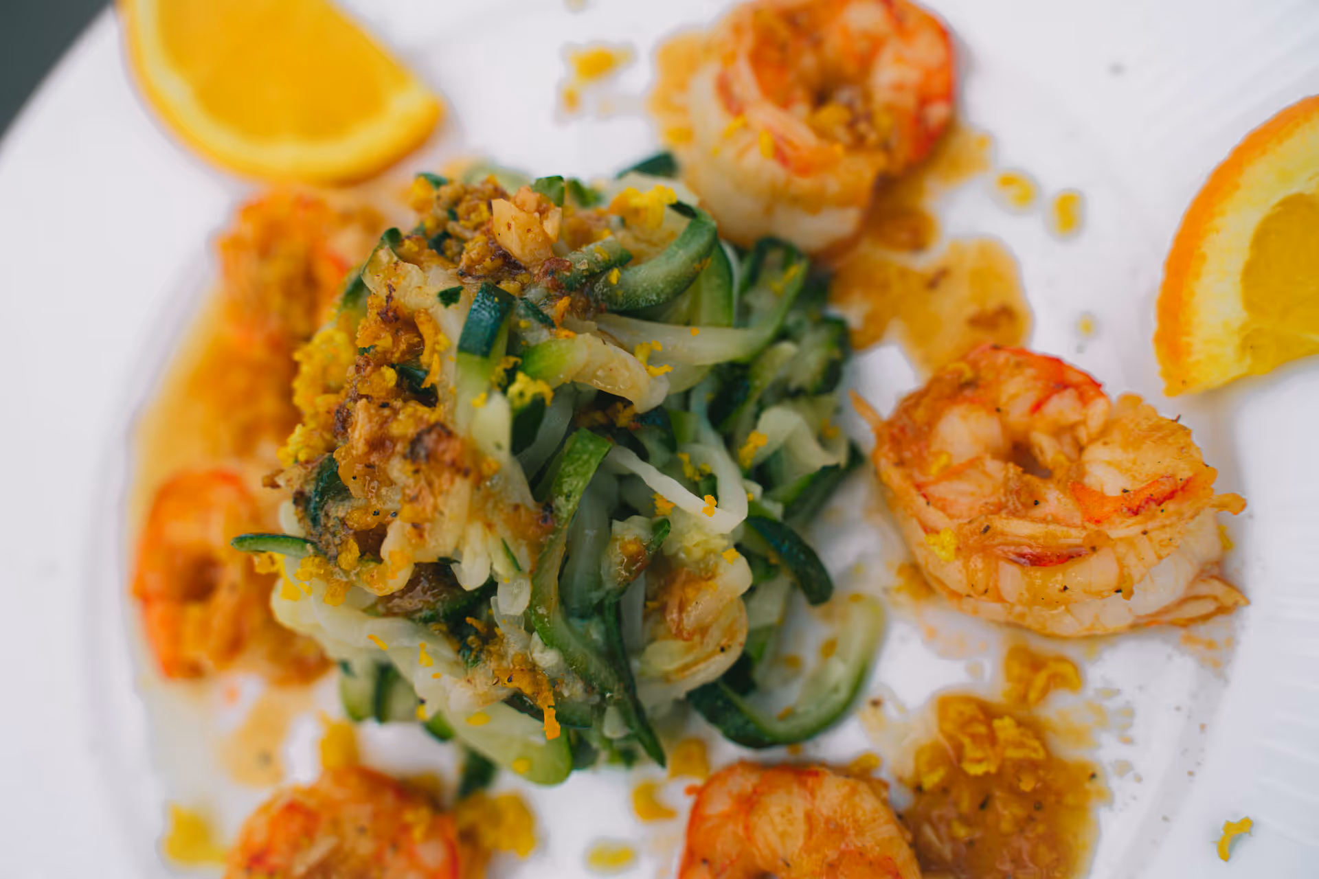 Close-up of a plate with cooked shrimp, spiralized zucchini, and garnished with orange slices and seasoning.