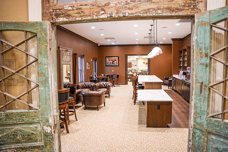View through rustic double doors into a warm communal dining and lounge area with leather seating, high-top tables, and a kitchen island with pendant lights.