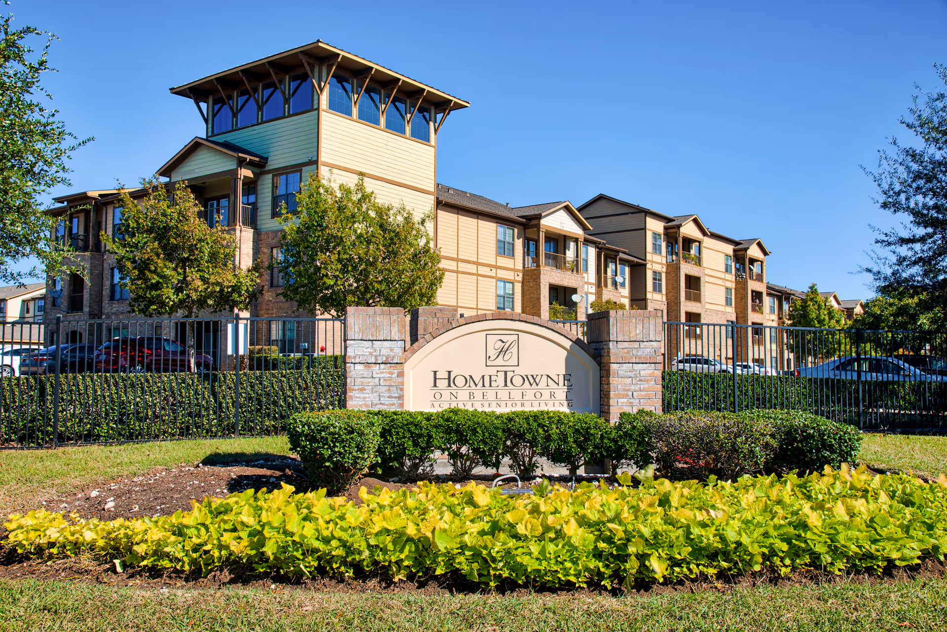 Exterior view of HomeTowne on Bellfort, an active senior living facility, showing a multi-story building with balconies, surrounded by trees and greenery, with a clear blue sky in the background. In front is a sign with the facility's name and a landscaped area with bushes and plants.