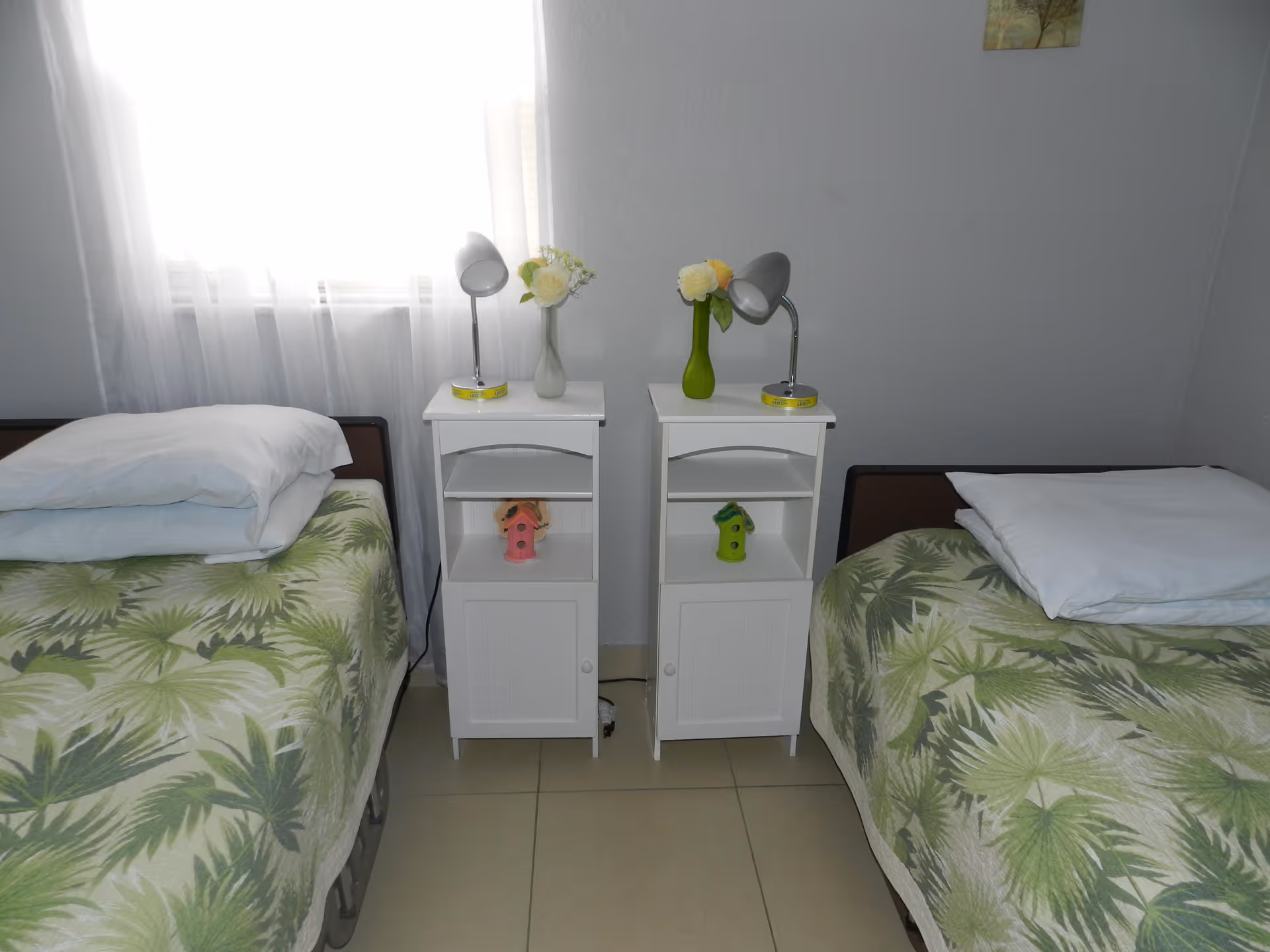 Two twin beds with green palm-patterned bedspreads and white nightstands with lamps and vases between them in a simple bedroom.