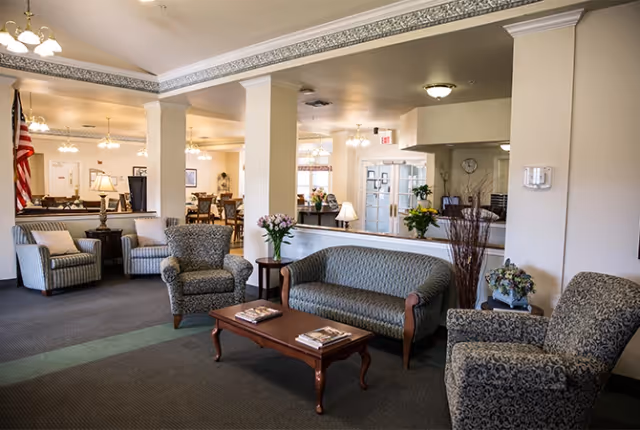 Spacious senior living common area with upholstered sofas and armchairs arranged around a wooden coffee table and a dining area visible in the background.