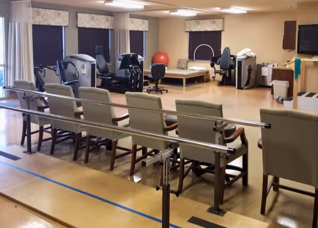 A rehabilitation room with parallel bars for walking exercises, several chairs lined up behind the bars, exercise equipment including a stationary bike and a leg press machine, a therapy bed, and a large red exercise ball. The room has windows with blinds and light-colored walls and flooring.