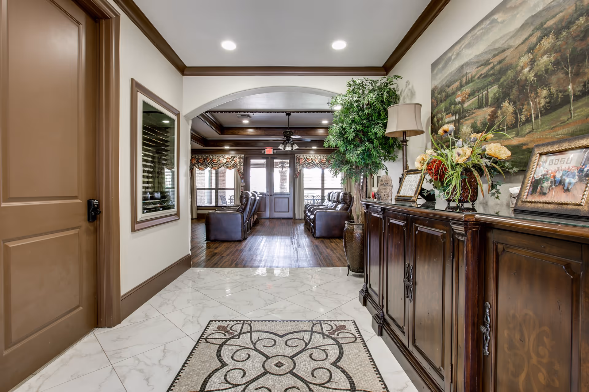 Interior view of a senior living facility hallway leading into a lounge area with leather recliners. The hallway has white marble tile flooring with a decorative mosaic rug, a wooden cabinet with framed photos and a flower arrangement, a floor lamp, and a large painting on the wall. The lounge area has wood flooring, large windows with patterned curtains, and ceiling fans with lights.