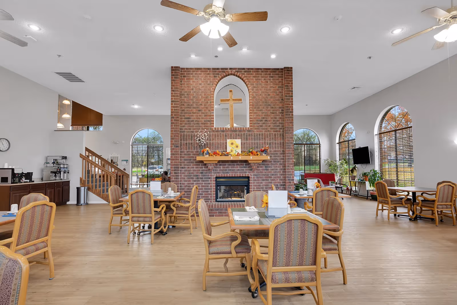 Spacious communal dining room with multiple tables and chairs centered around a large brick fireplace, arched windows, and ceiling fans.