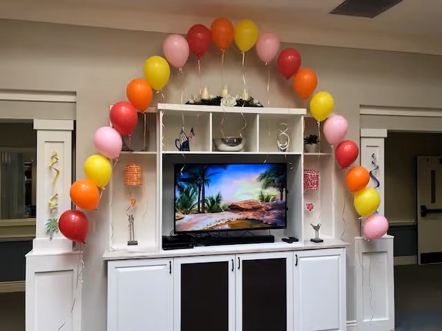 A white entertainment center with a flat-screen TV displaying a tropical beach scene. The unit has shelves with decorative items and two small American flags. Colorful balloons in red, orange, yellow, and pink are arranged in an arch around the entertainment center. The setting appears to be an indoor common area with neutral-colored walls and doors on either side.