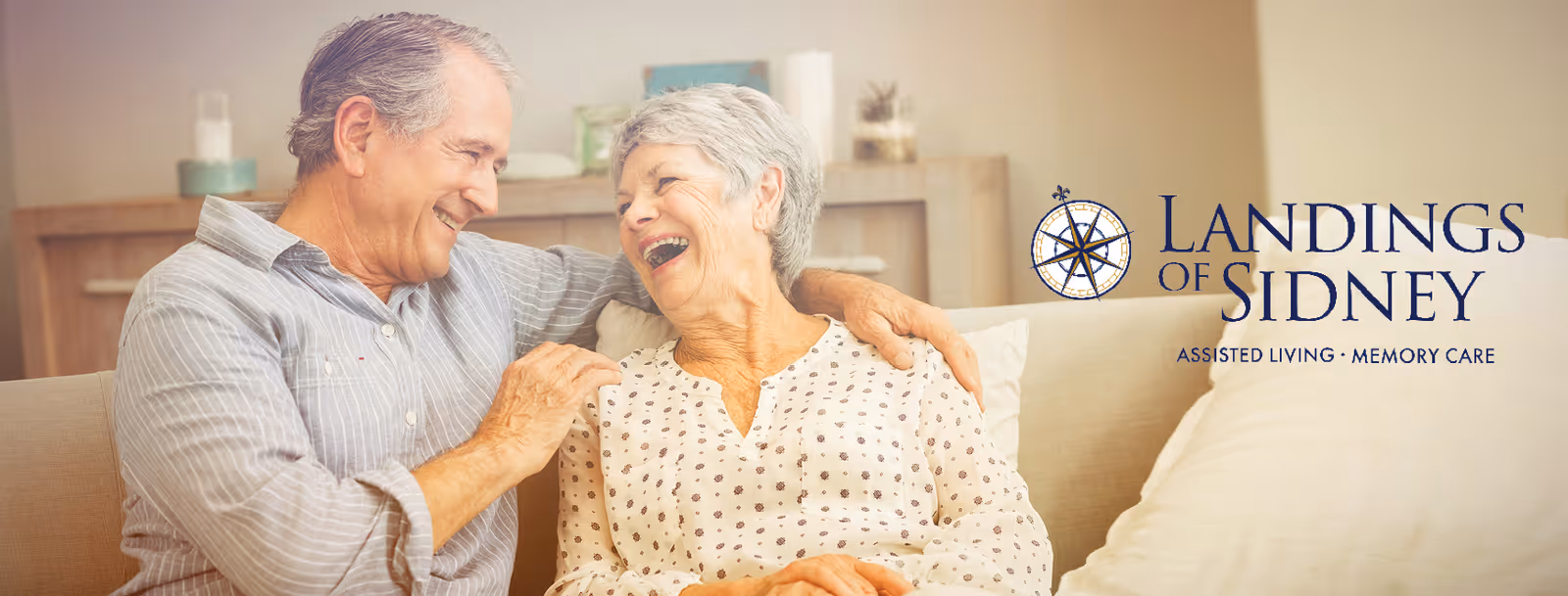 An elderly couple sitting on a beige couch in a cozy living room. The man is smiling and has his arm around the woman, who is laughing joyfully. Behind them is a wooden shelf with decorative items. The logo for Landings of Sidney, an assisted living and memory care facility, is displayed on the right side of the image.