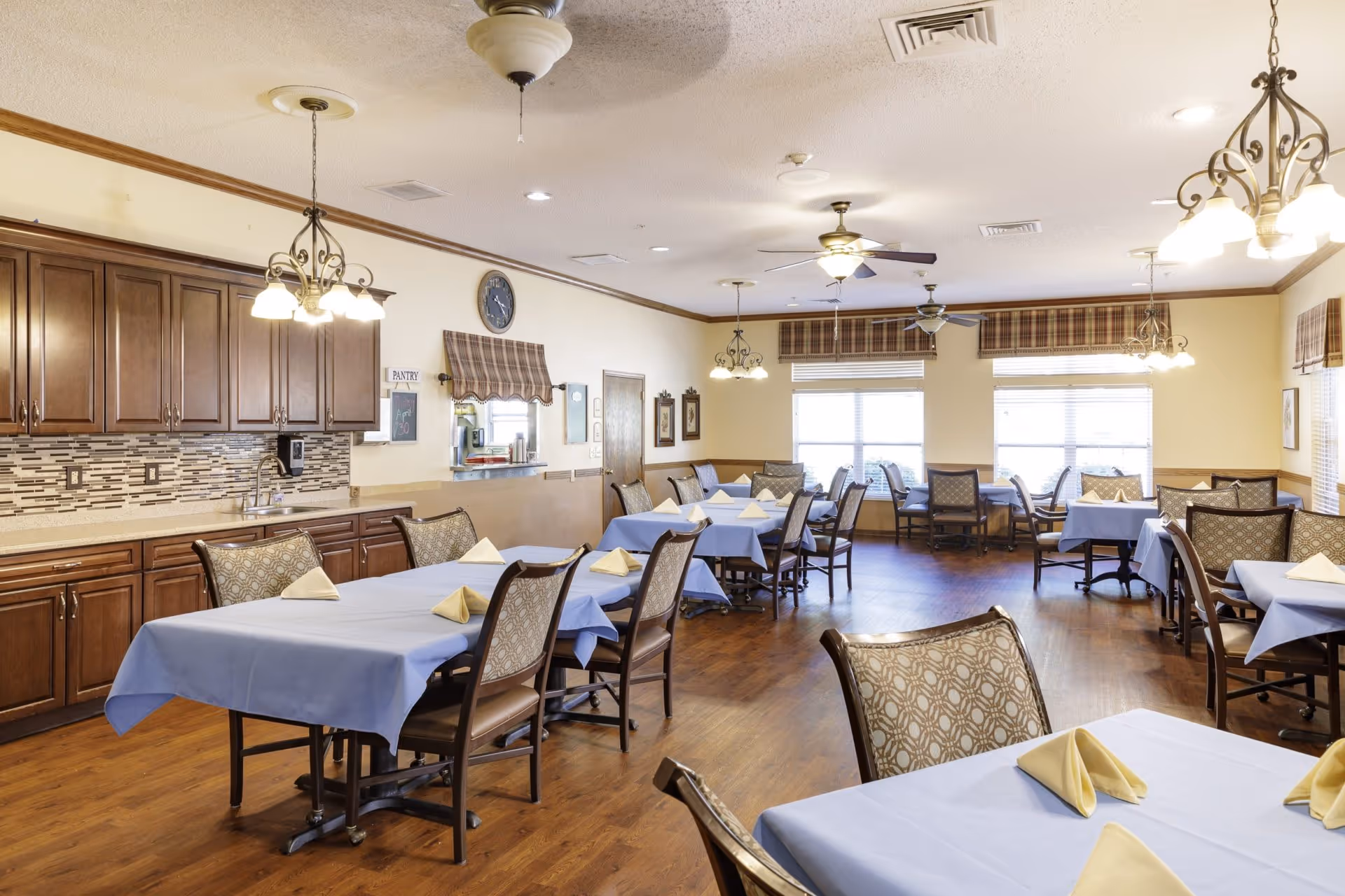 Bright dining room with multiple tables covered in light blue tablecloths and folded napkins, wooden chairs, cabinets, and large windows.