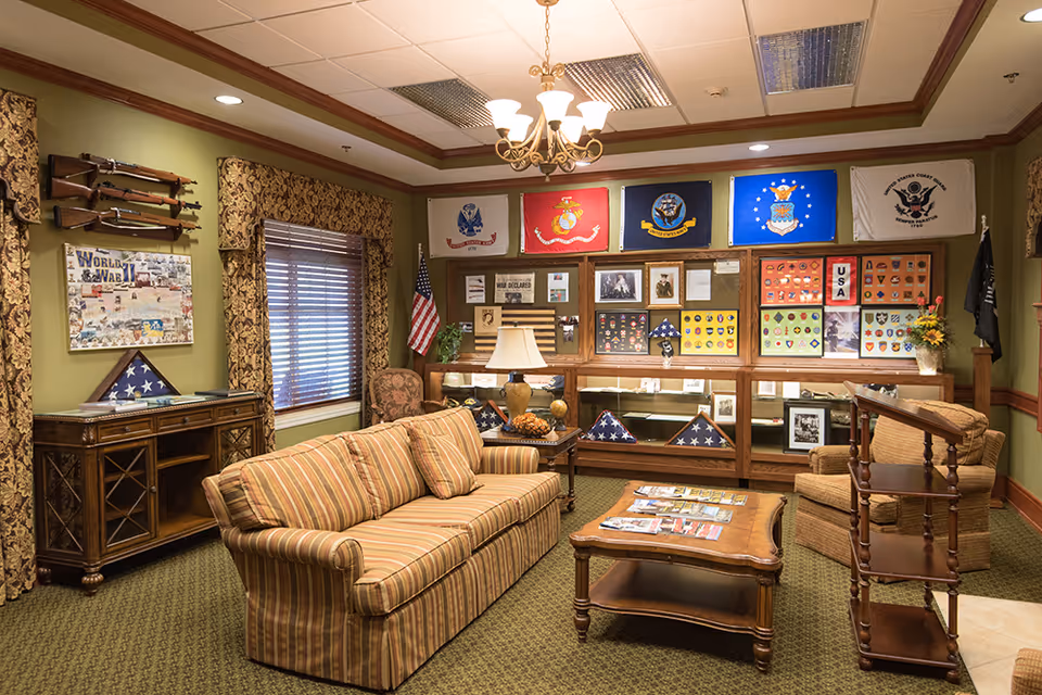 A cozy living room with a striped sofa, armchair, wooden coffee table, and side tables. The walls are decorated with military memorabilia including flags, framed photos, medals, and rifles mounted on the wall. There is a window with patterned curtains and a chandelier hanging from the ceiling.