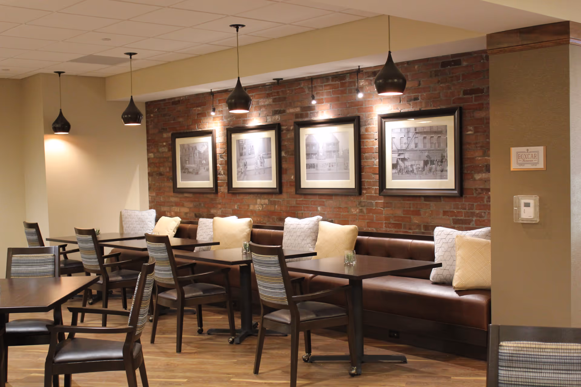 Cozy dining area with tables, chairs and banquette seating along a brick wall adorned with framed pictures and pendant lights.
