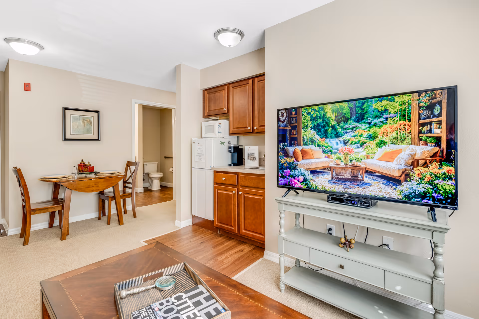 Interior view of a senior living facility room featuring a wooden dining table with two chairs, a kitchenette with wooden cabinets, a microwave, and a coffee maker. A large flat-screen TV on a white stand displays a vibrant garden scene. In the background, a bathroom with a visible toilet is seen through an open door.