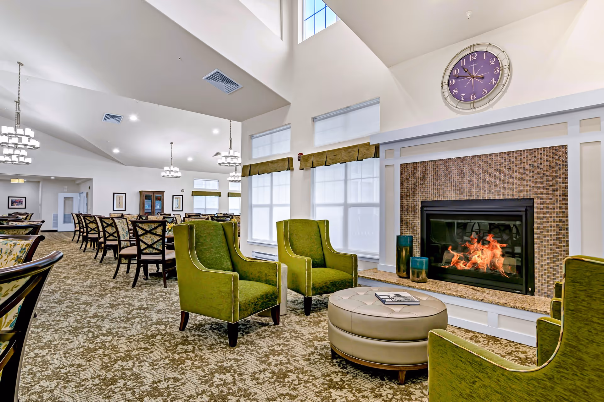 A spacious and well-lit common area in a retirement living facility featuring a cozy fireplace with a mosaic tile surround, a large purple clock above it, and three green upholstered armchairs arranged around a round beige ottoman. In the background, there are multiple dining tables with chairs, decorative light fixtures hanging from the ceiling, and large windows with white shades and green valances.