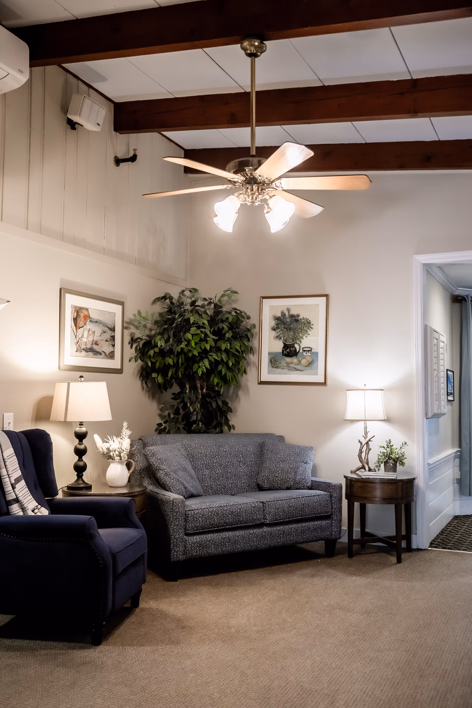 Cozy seating area with a loveseat, armchair, side tables and lamps, wall art, a potted plant, and a ceiling fan in a common living room.