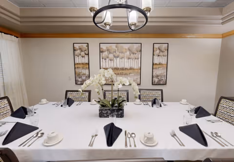 A formal dining room with a large rectangular table covered with a white tablecloth. The table is set with black folded napkins, white cups and saucers, glasses, and silverware. A centerpiece with white orchids is placed in the middle of the table. The room has beige walls with three framed paintings of trees hanging on the wall and a modern chandelier hanging from the ceiling.