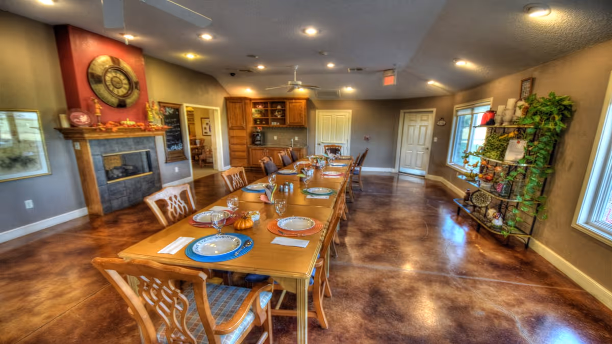 A spacious dining room with a long wooden table set with plates, glasses, and napkins. The room features a fireplace with a large decorative clock above it, wooden chairs with patterned cushions, a shelving unit with plants and decorative items, and warm lighting from recessed ceiling lights.