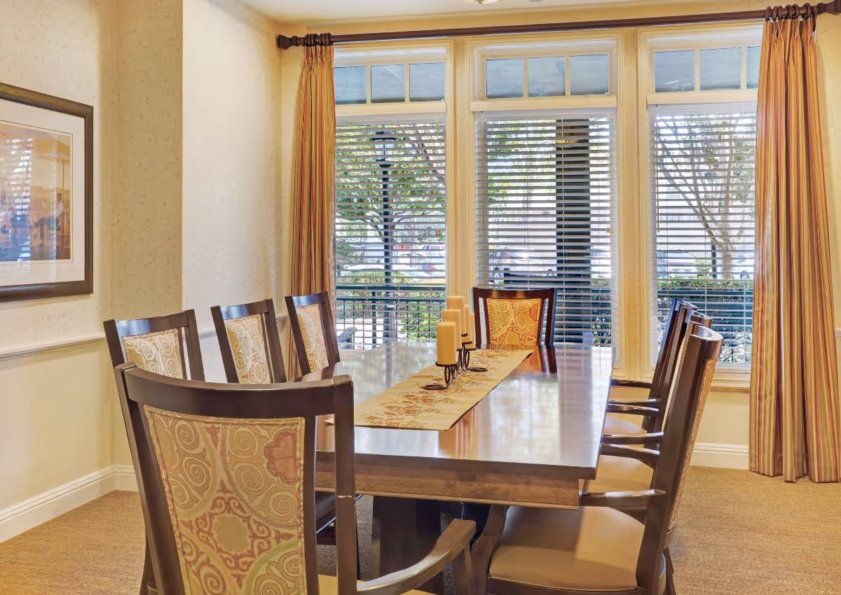 A dining room with a long wooden table surrounded by eight chairs with patterned upholstery. The table has a decorative runner and a candle holder with multiple candles. Large windows with blinds and striped curtains allow natural light to enter the room.